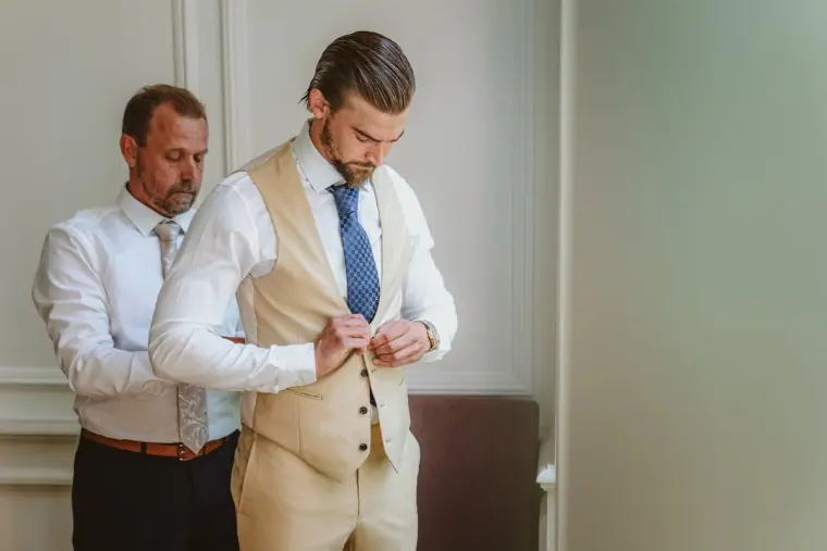 Groom buttoning his beige waistcoat as an older man adjusts his collar