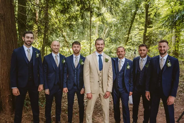 Groom in a beige suit with groomsmen in navy suits standing among forest trees