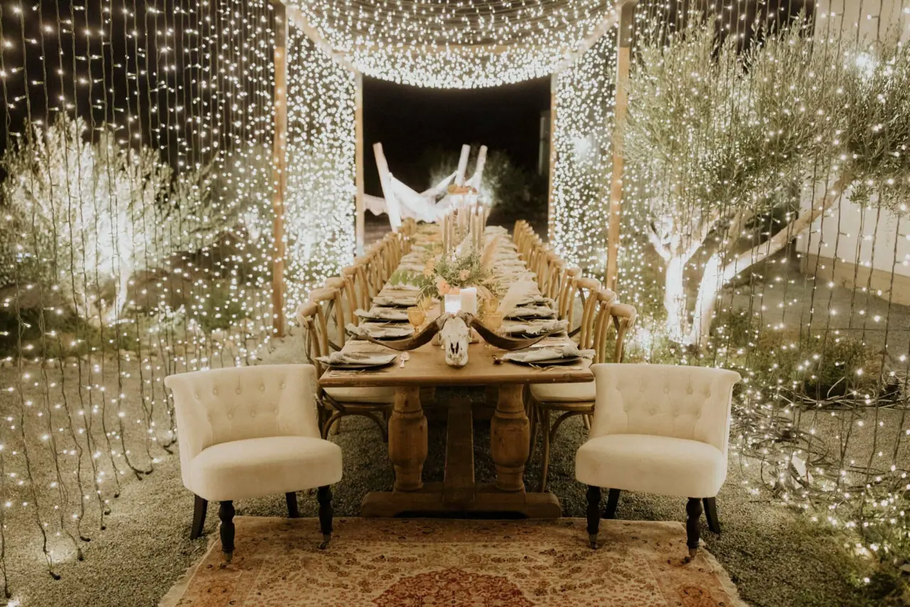 Long banquet table with cream armchairs under curtains of fairy lights and white branches at Aelia