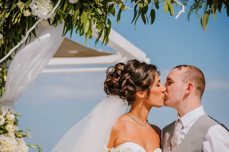 Louise and Mike kissing beneath the greenery-draped arch against a blue sky