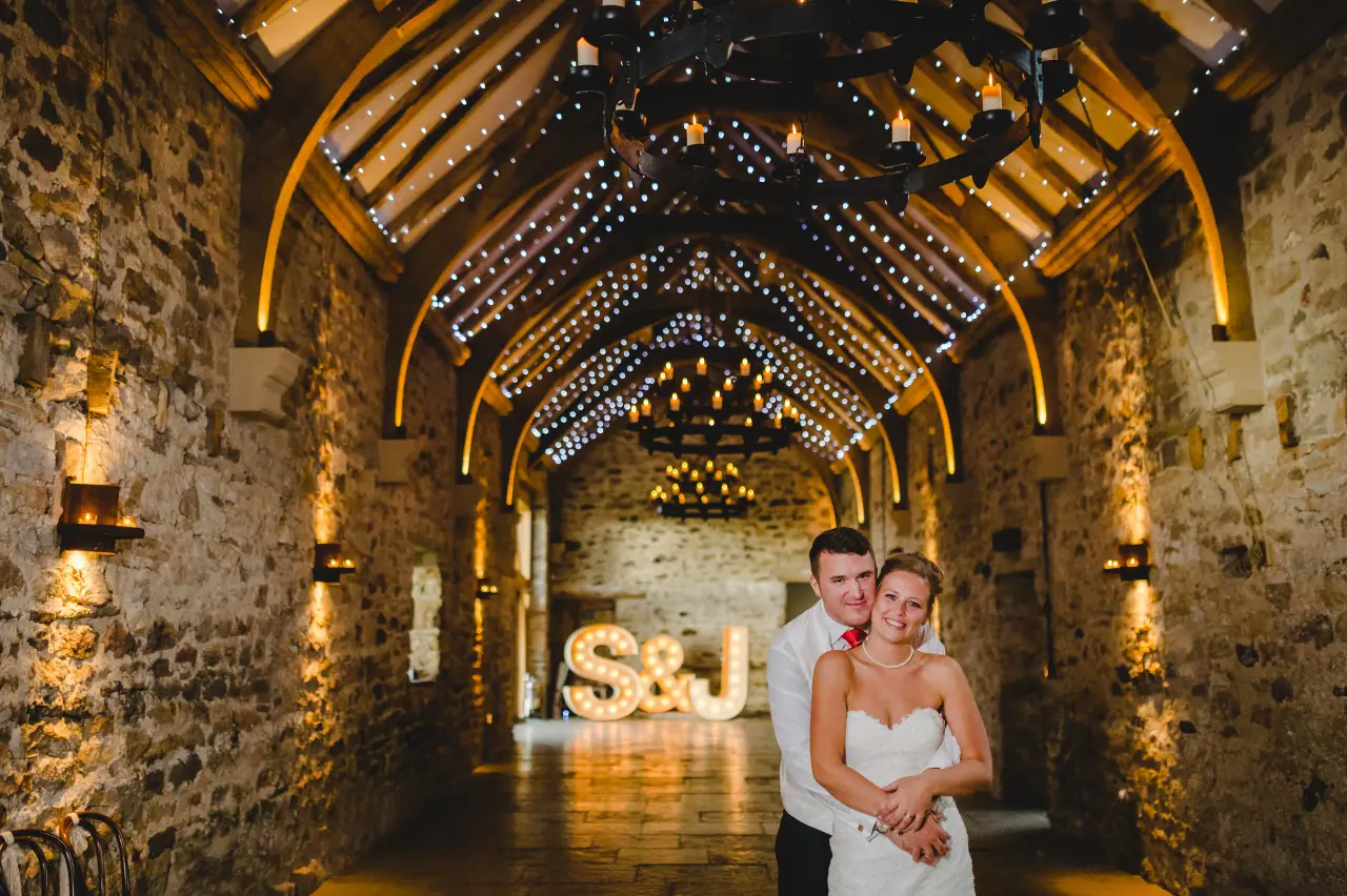 Sonny and Jo embracing in the fairy-lit stone hall at Healey Barn, Northumberland, with S&J light-up letters