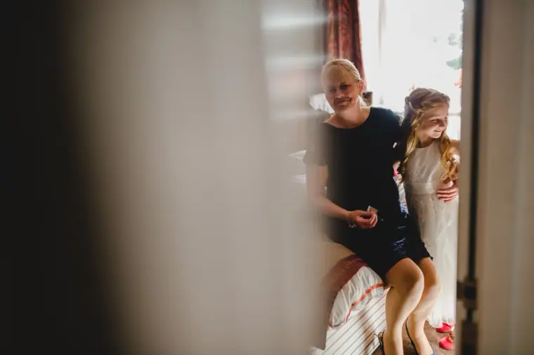 Bride hugging a young bridesmaid by a window, seen through a doorway