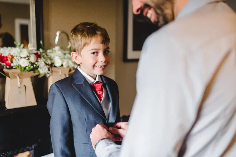 Young pageboy in a navy suit and red tie having his jacket adjusted