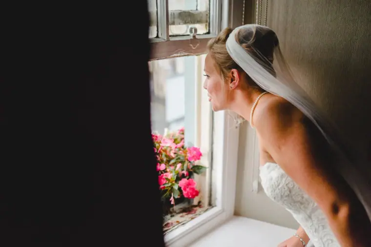 Bride in her veil gazing out of a window with pink flowers on the sill