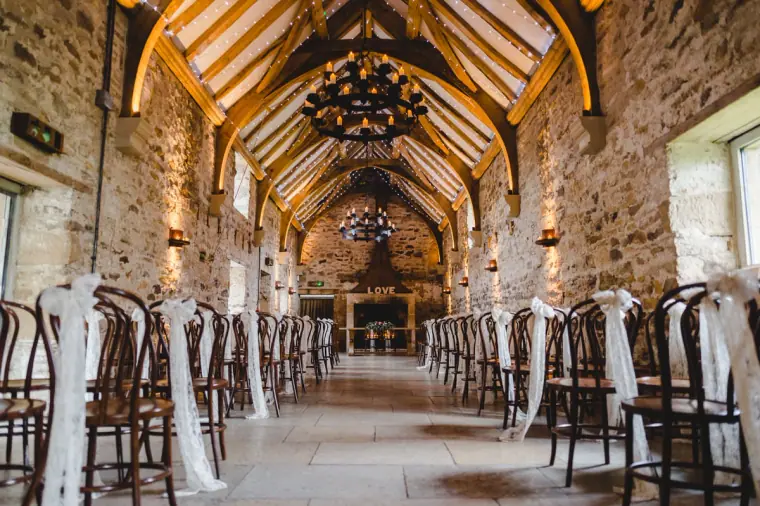 Ceremony hall at Healey Barn with exposed beams, chandeliers and chairs tied with white ribbon