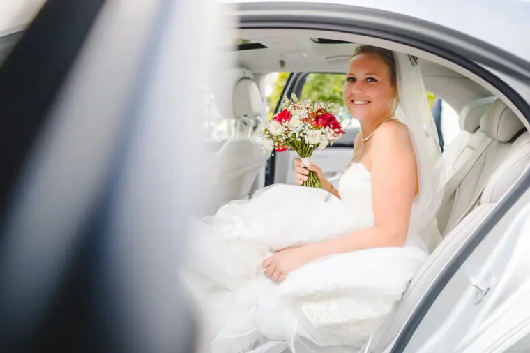 Bride smiling in the back of the wedding car with her red and white bouquet