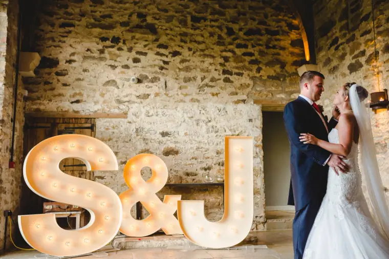 Sonny and Jo embracing beside the glowing S&J letters against the stone wall
