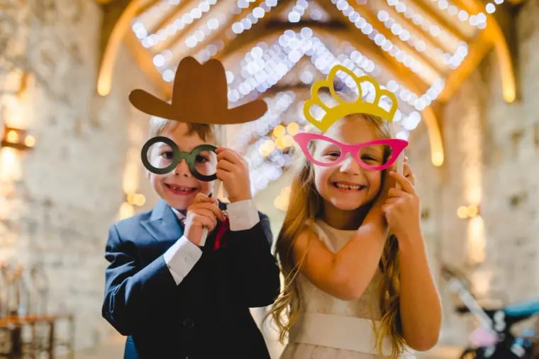 Young pageboy and bridesmaid posing with photo-booth glasses, hat and crown props