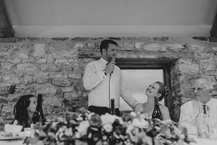 Black-and-white photo of the groom giving his speech as the bride laughs