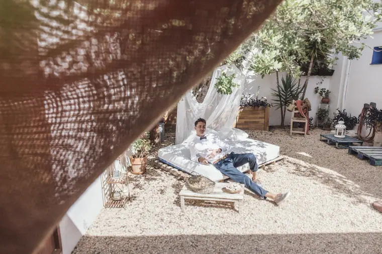 Groom lounging on a daybed in a white-walled courtyard, seen past hessian fabric