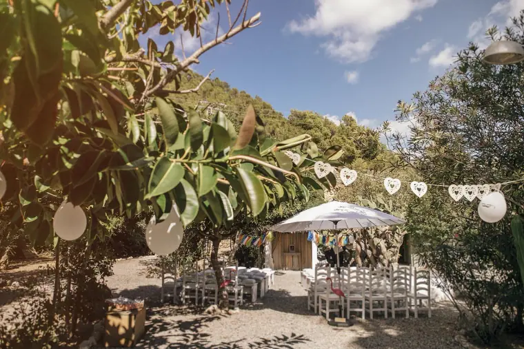 Ceremony chairs set beneath a white umbrella among trees, with hanging lanterns