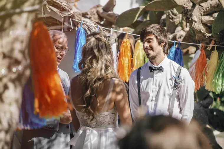 Bride and groom standing before the celebrant beneath colourful tassel garlands