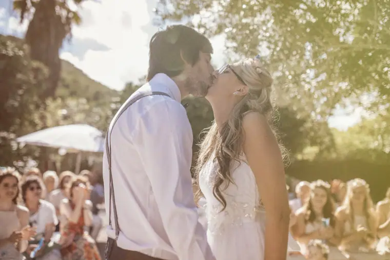 Bride and groom's first kiss in dappled sunlight as guests look on