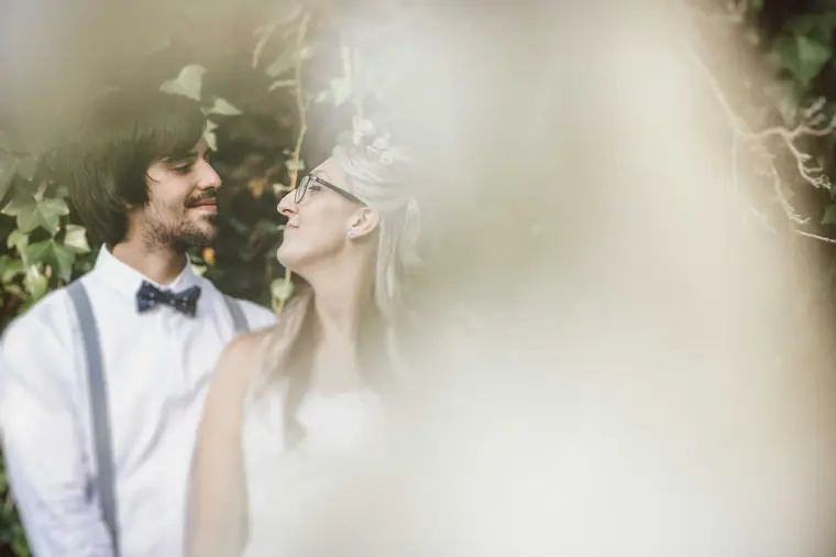 Groom in a bow tie smiling at the bride through soft, hazy foliage