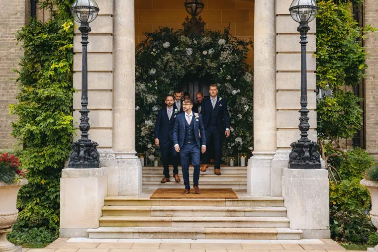 Ian and groomsmen in navy suits walking down the steps of the floral entrance