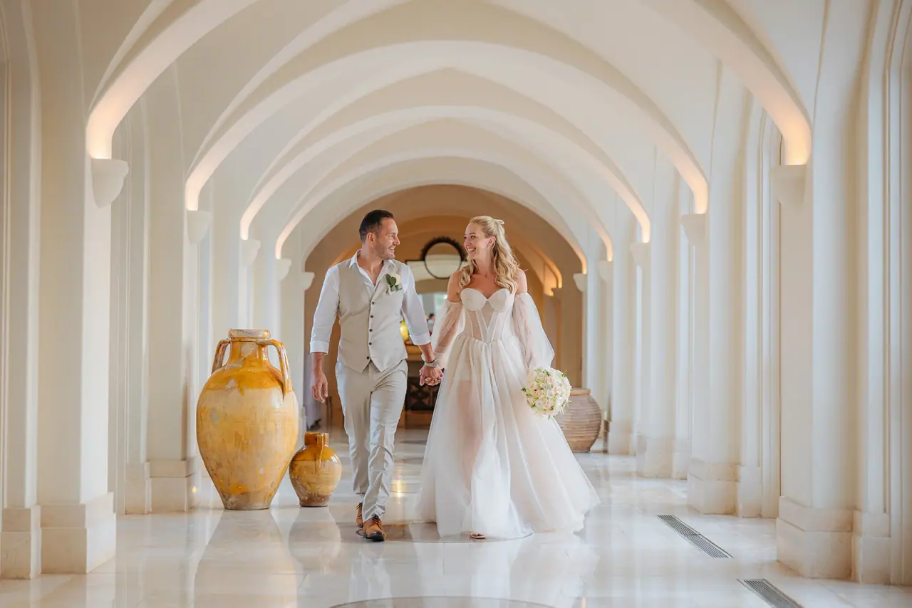 Jade and Peter walking hand in hand through the white arched colonnade at Anassa Hotel