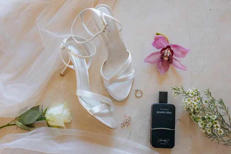Bridal sandals, rings, a pink orchid and cologne laid out on cream fabric