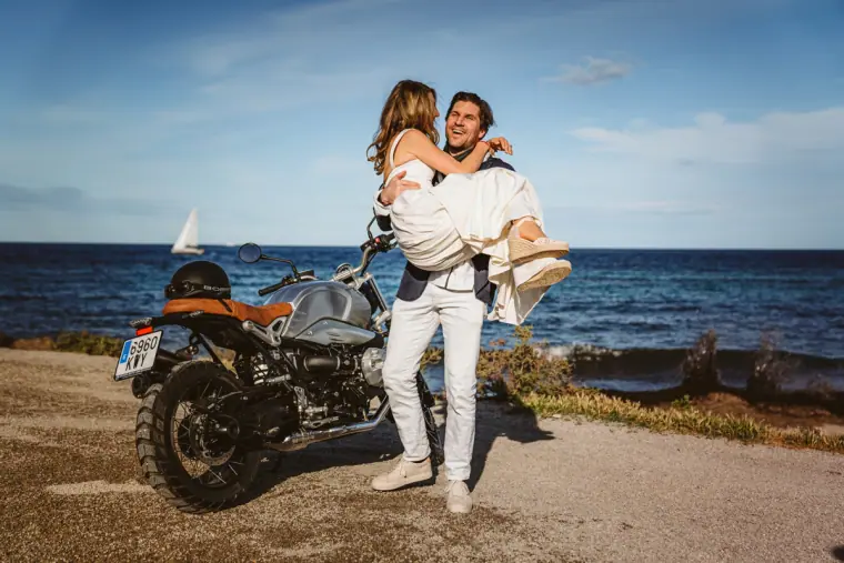 Antonia and Birger laughing as he carries her beside a vintage motorbike above the Ibiza coastline