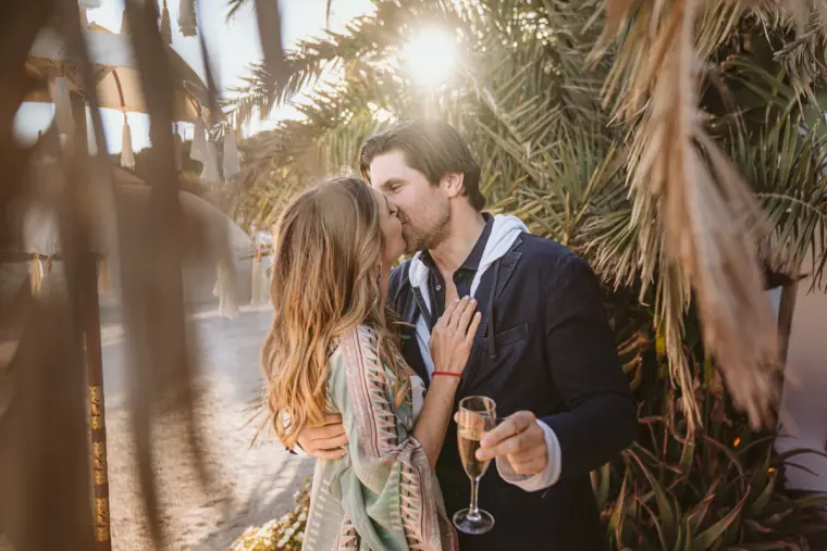 Couple kissing among palm leaves in golden backlight, glass of champagne in hand