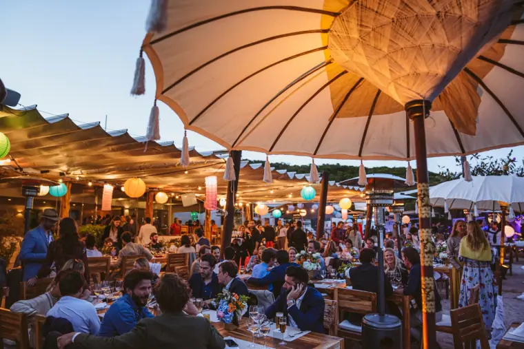 Packed terrace of guests dining under parasols and glowing lanterns at dusk at La Escollera