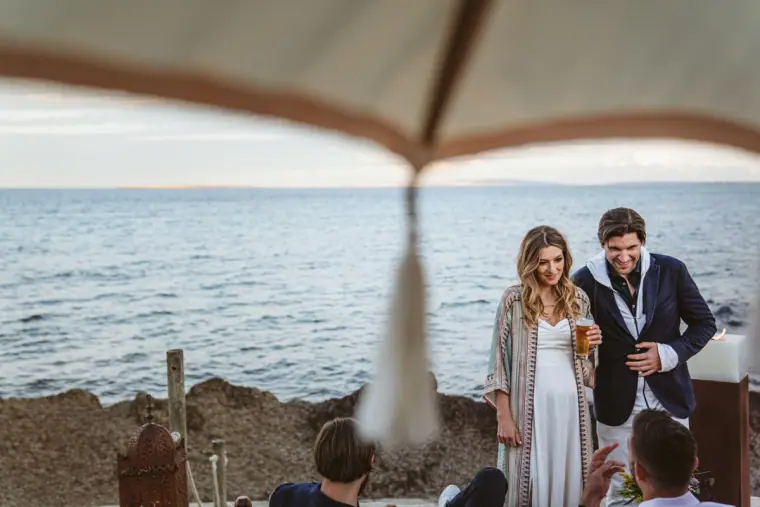 Couple chatting with guests on the rocks by the sea, framed by a parasol fringe