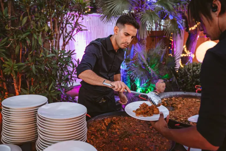 Chef serving a giant pan of paella beside stacks of plates under purple lights