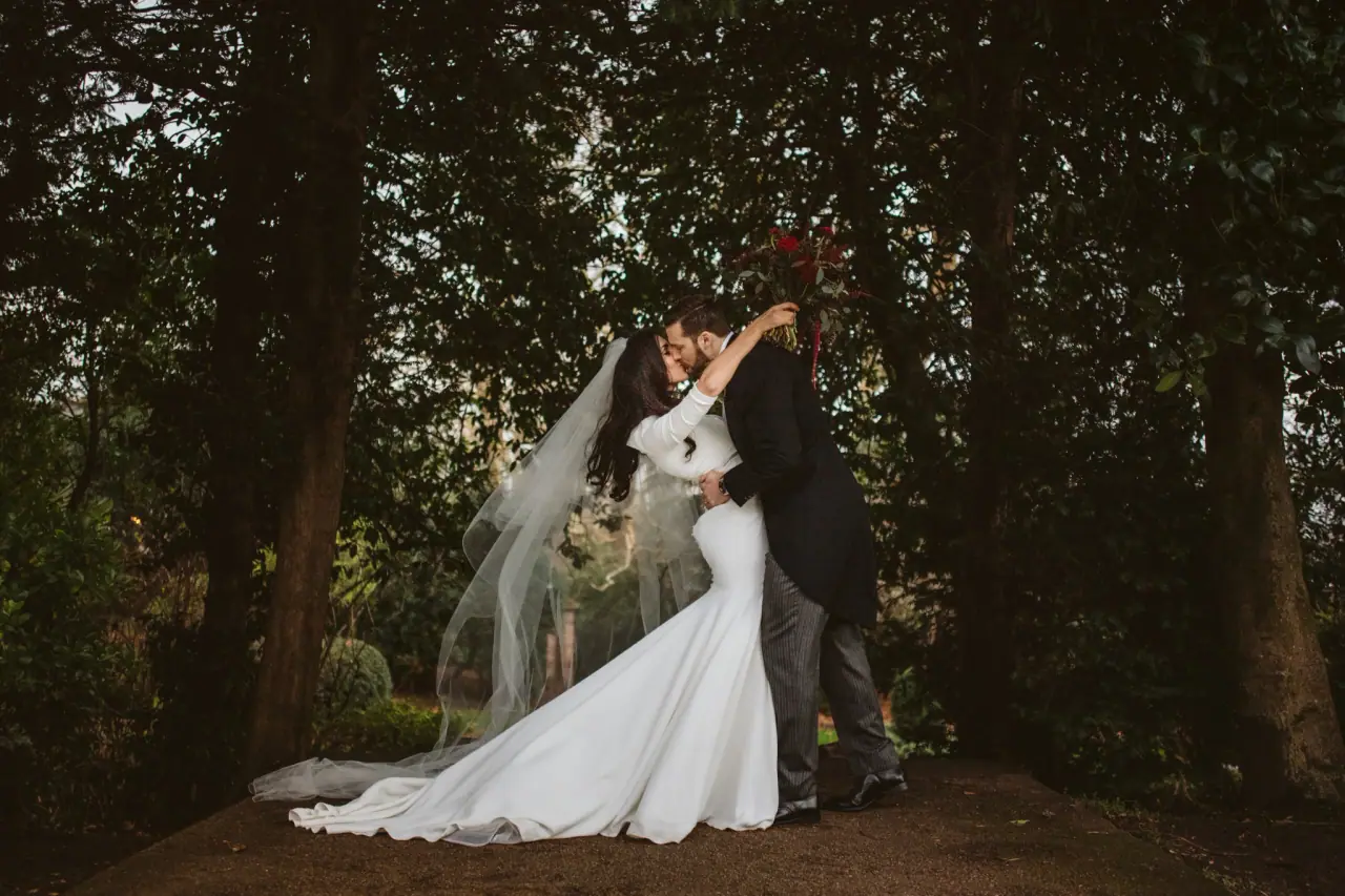 Stephanie and Jamie kissing among dark trees, her long veil and train spread on the ground, London