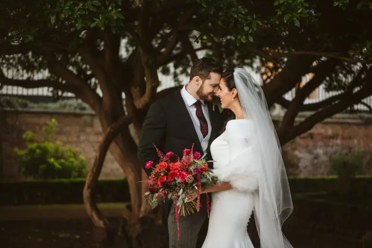 Groom kissing the bride's forehead as she holds a red winter bouquet under a tree