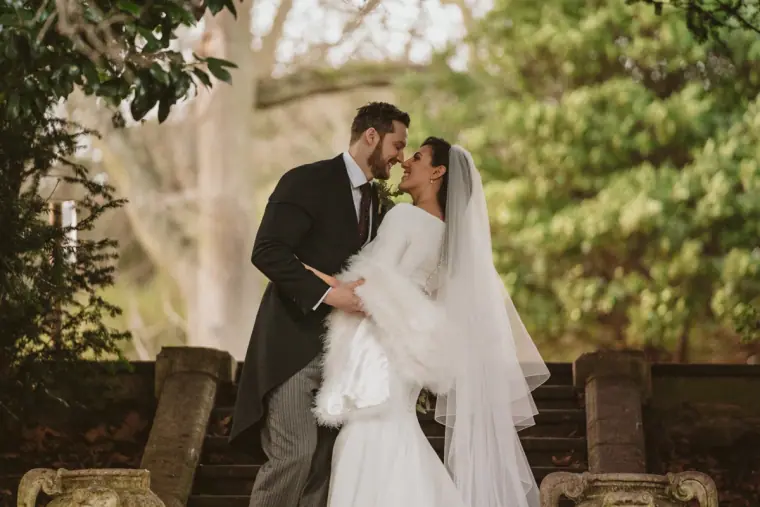 Stephanie and Jamie embracing on garden steps, the bride in a white fur stole and long veil