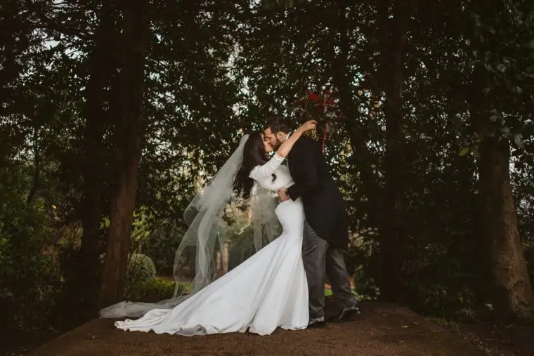 Bride and groom kissing beneath tall trees, her cathedral veil trailing, before The Landmark reception