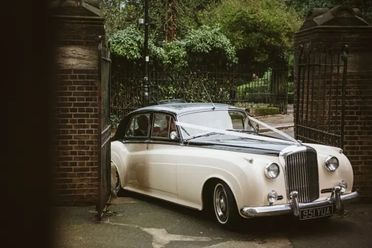 Vintage ivory Bentley wedding car with ribbons parked by wrought-iron gates