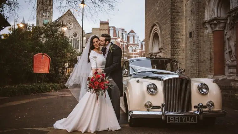 Bride with red bouquet and groom beside the vintage ivory Bentley outside the church