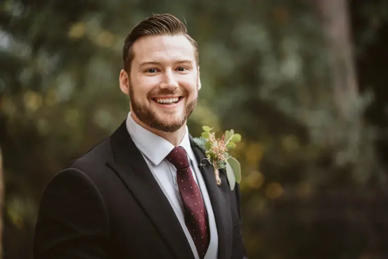 Smiling groom in a dark suit, burgundy tie and winter buttonhole