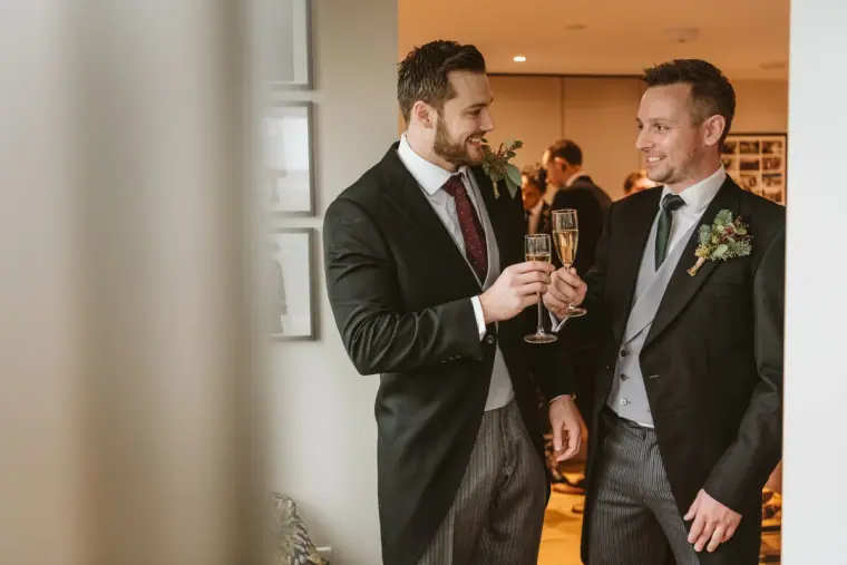 Groom and best man raising champagne glasses at home before the wedding
