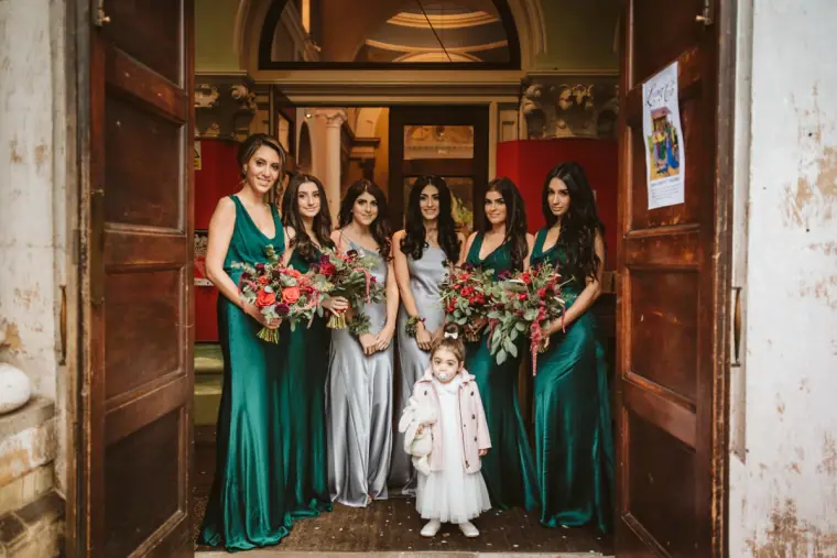 Bridesmaids in emerald and silver gowns with a flower girl in a church doorway
