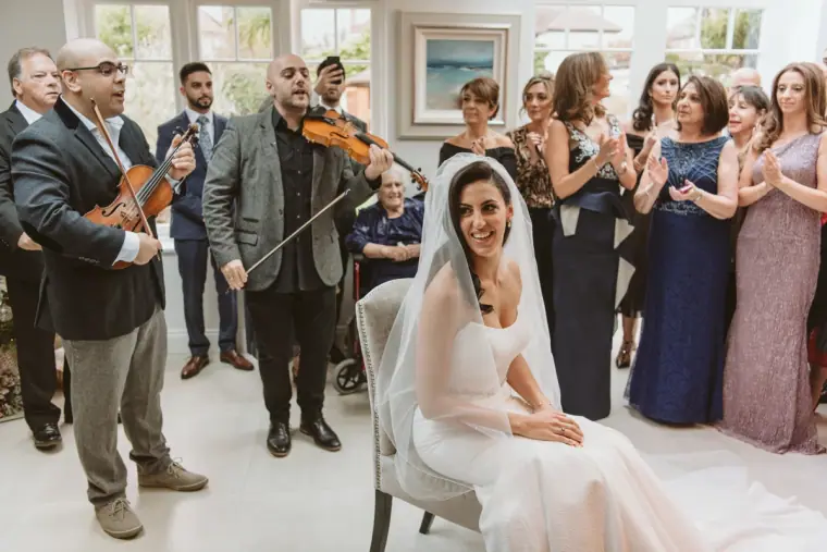 Musicians playing violins for the seated bride as family gather around at home