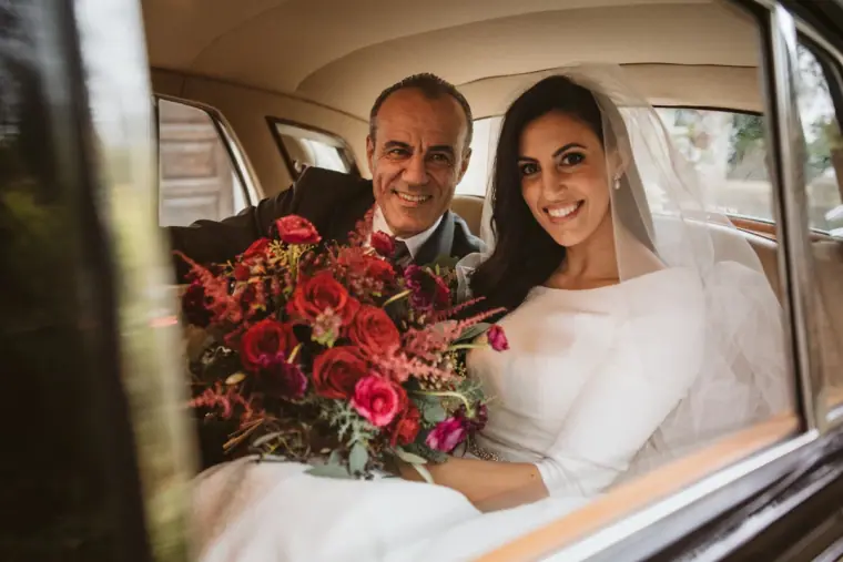 Smiling bride and her father with a red bouquet in the back of the wedding car