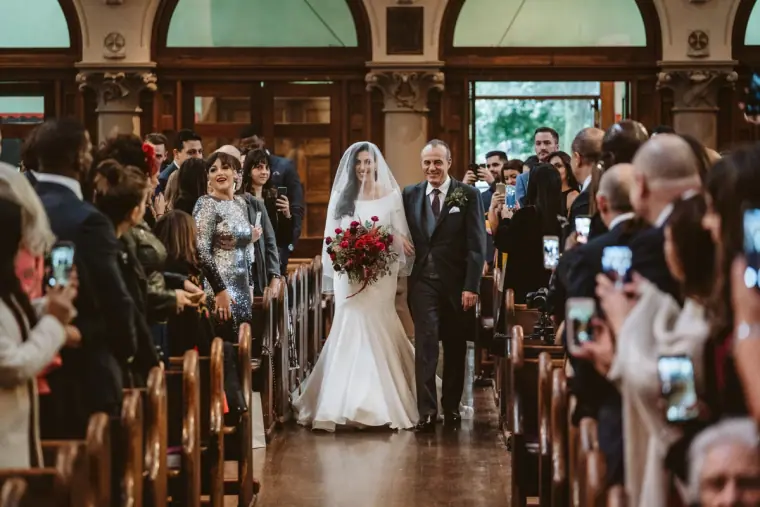 Bride walking down the aisle on her father's arm past guests holding up phones