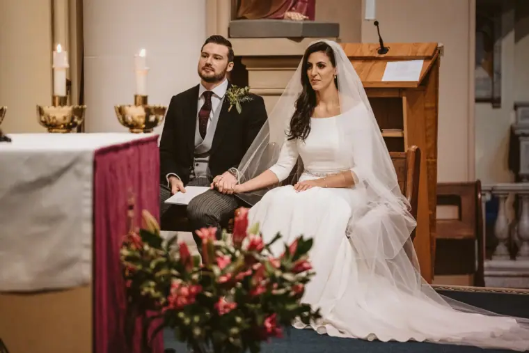 Bride and groom seated side by side during the ceremony, red flowers in front