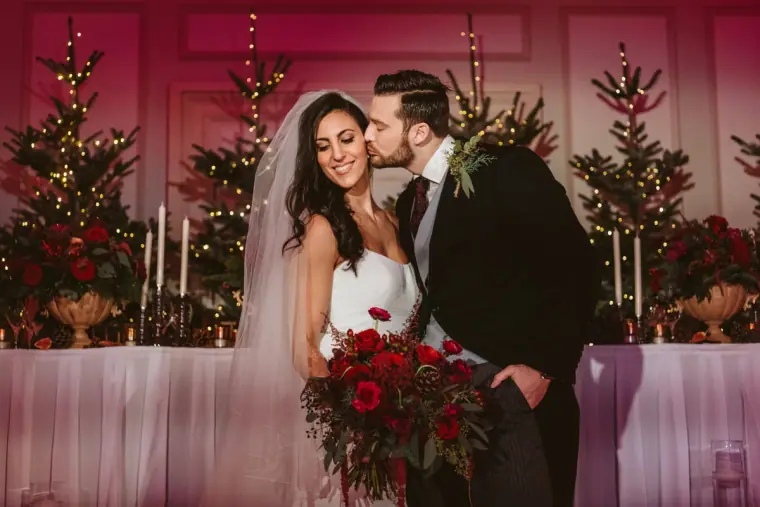 Couple kissing in front of red-draped tables and Christmas trees in the ballroom at The Landmark London