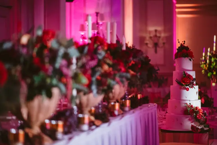 Red floral centrepieces, candles and a white tiered cake on the top table under pink light