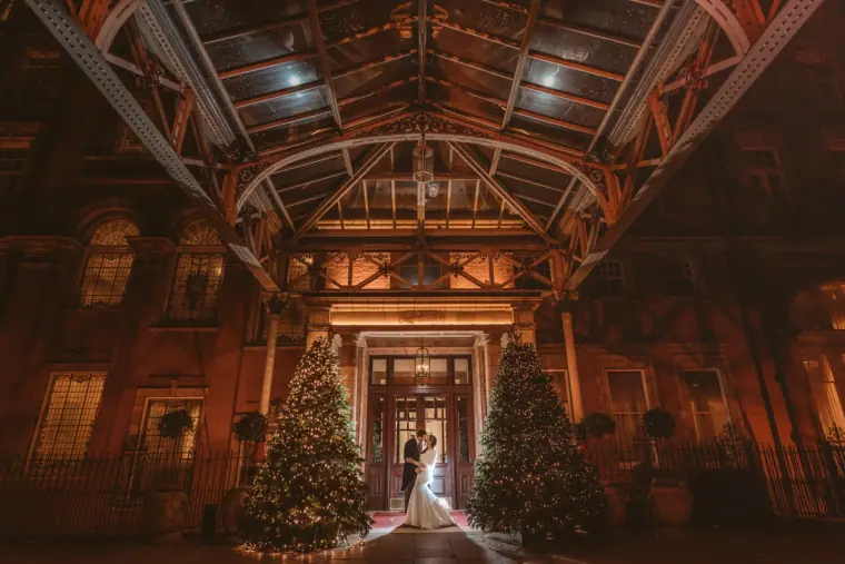 Couple framed by Christmas trees beneath the glass canopy of The Landmark London at night