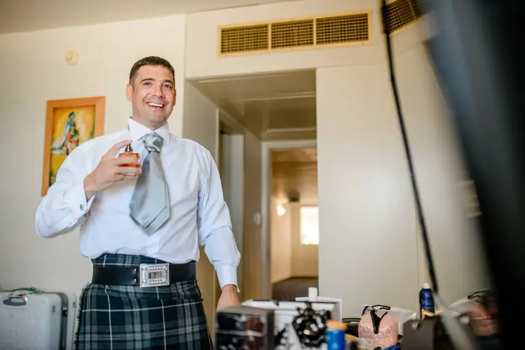 Man in a kilt and grey tie laughing with a drink while getting ready
