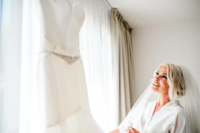 Bride in her robe and veil smiling up at her gown hanging by the window