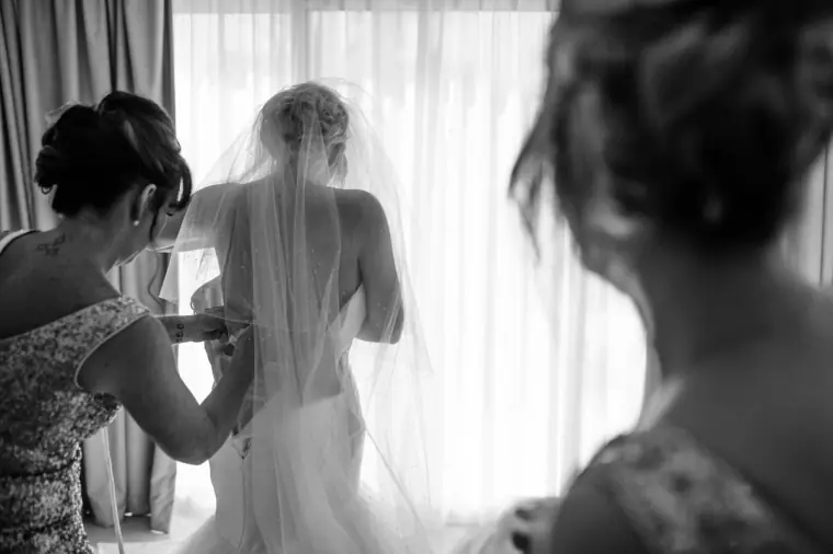 Black-and-white photo of bridesmaids lacing the back of the bride's gown by the window