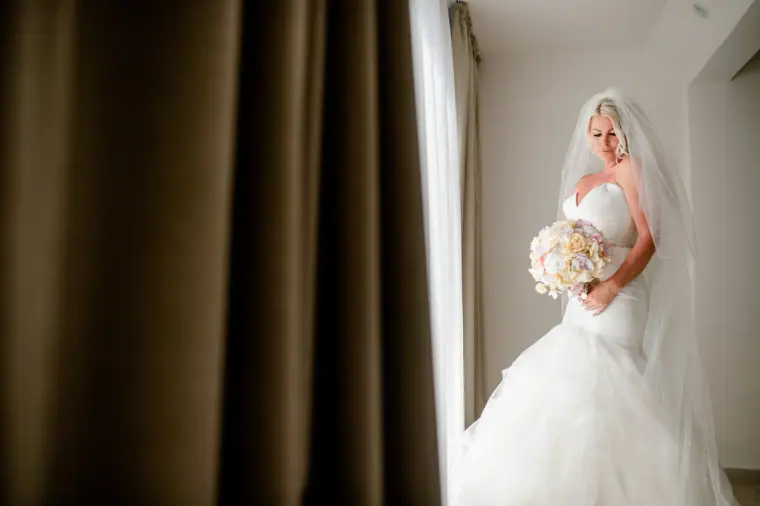 Bride holding her pastel bouquet in window light, framed by a dark curtain