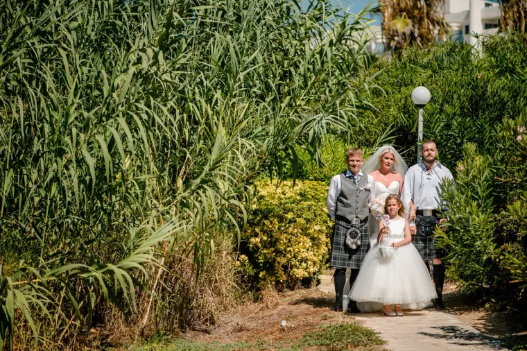 Bride walking down a green garden path with men in kilts and the flower girl
