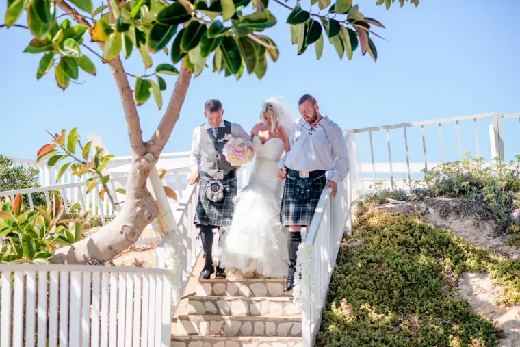 Bride escorted down white steps beneath a tree by two men in kilts