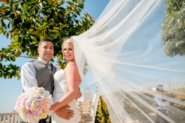 Bride's veil billowing in the breeze as she stands with the groom under trees
