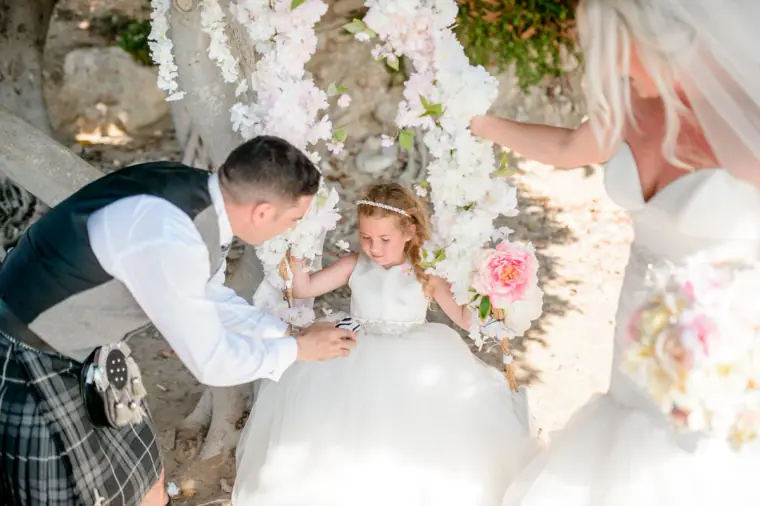 Groom kneeling to give the flower girl a gift beneath a floral arch